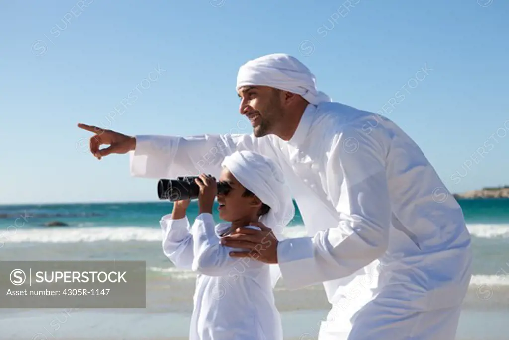 Arab father and son looking through binoculars by the beach.