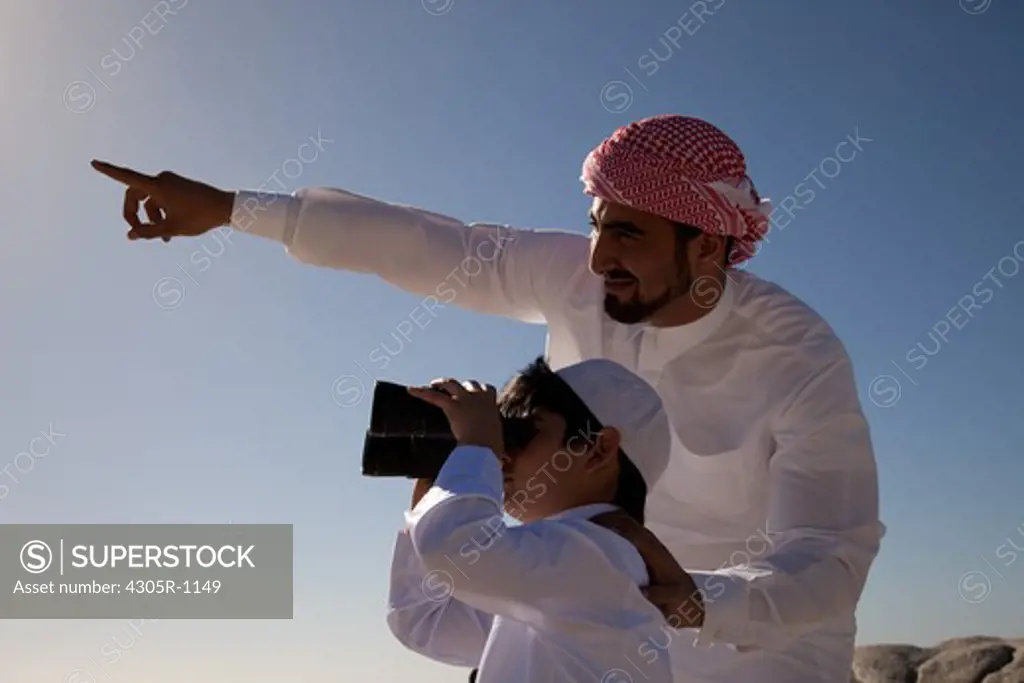 Arab father and son looking through binoculars by the beach.
