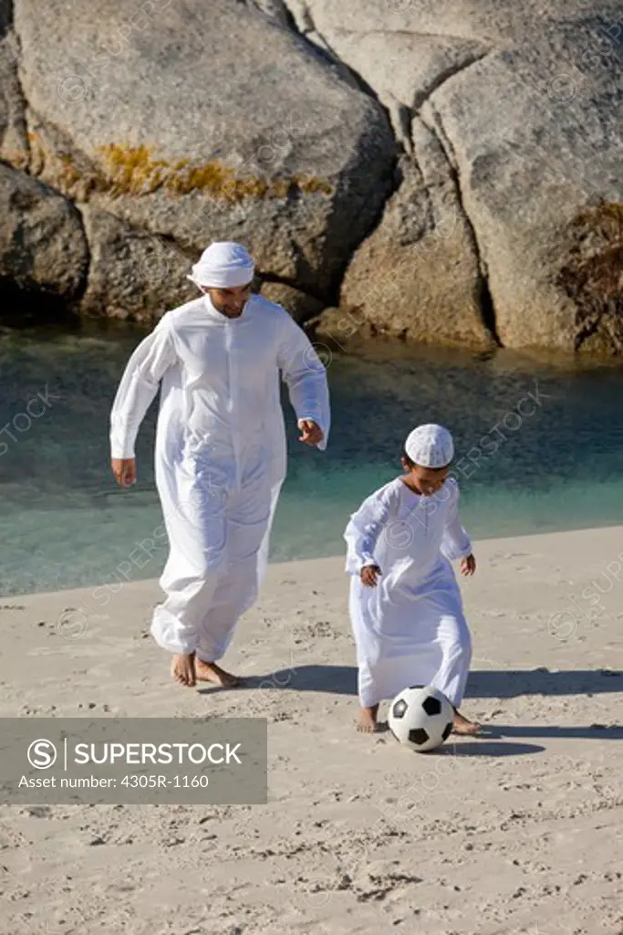 Arab father and son playing soccer ball by the beach.