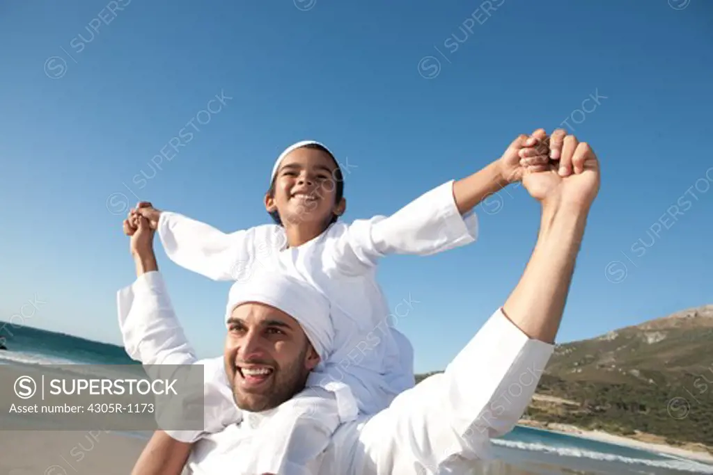 Arab father carrying his son on shoulders at the beach, smiling.