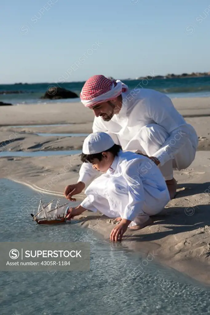 Arab father and son playing toy boat on beach.