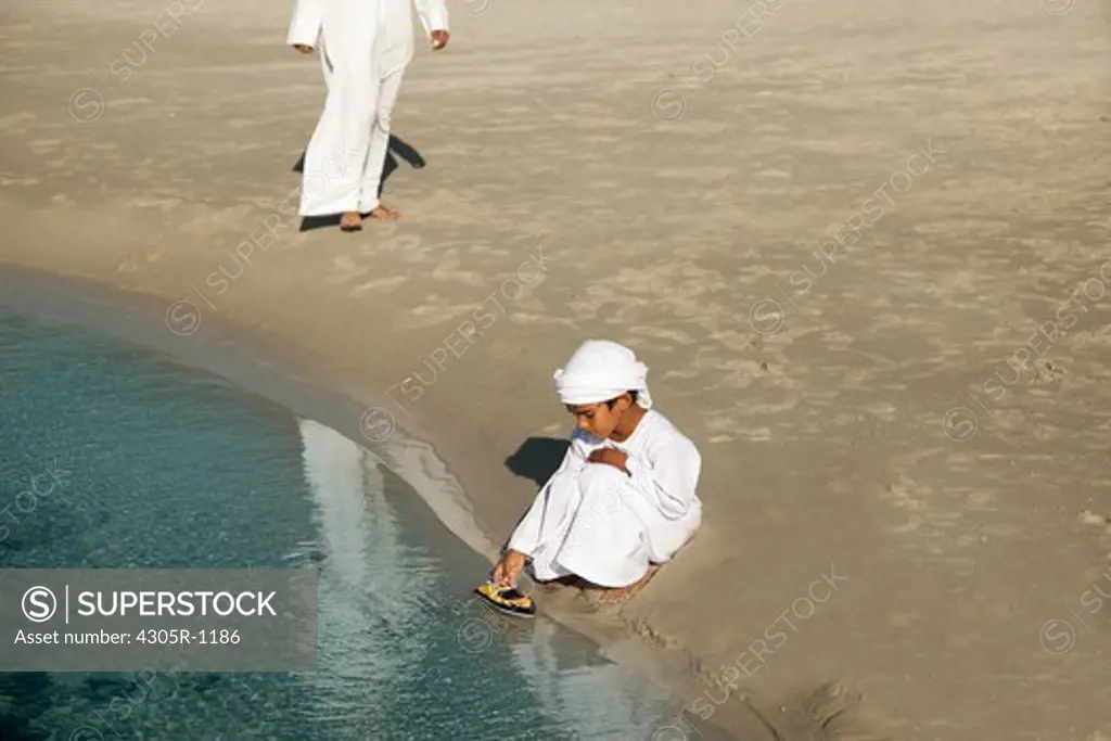 Arab boy playing toy boat at the beach, man walking towards the boy.