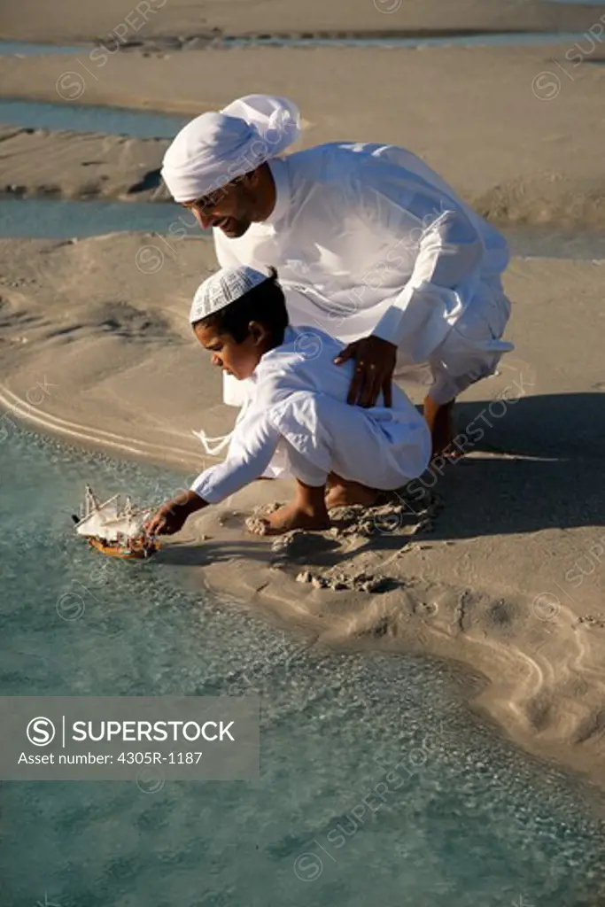 Arab father and son playing toy boat on beach.