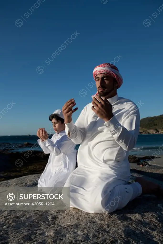 Arab father and son praying, kneeling on beach.