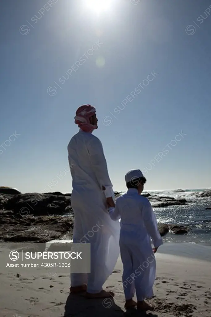 Arab father and son standing by the beach, facing the sea.