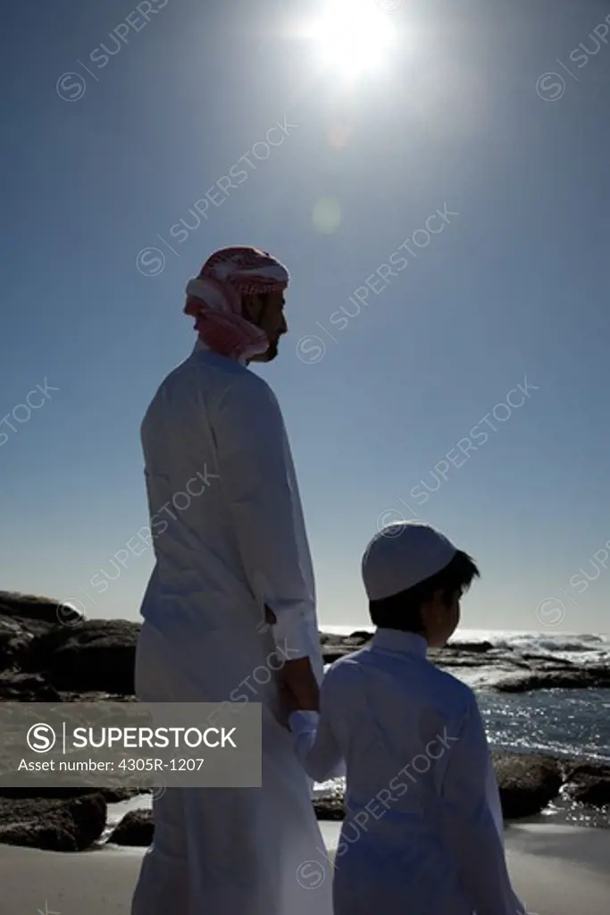 Arab father and son standing by the beach, facing the sea.