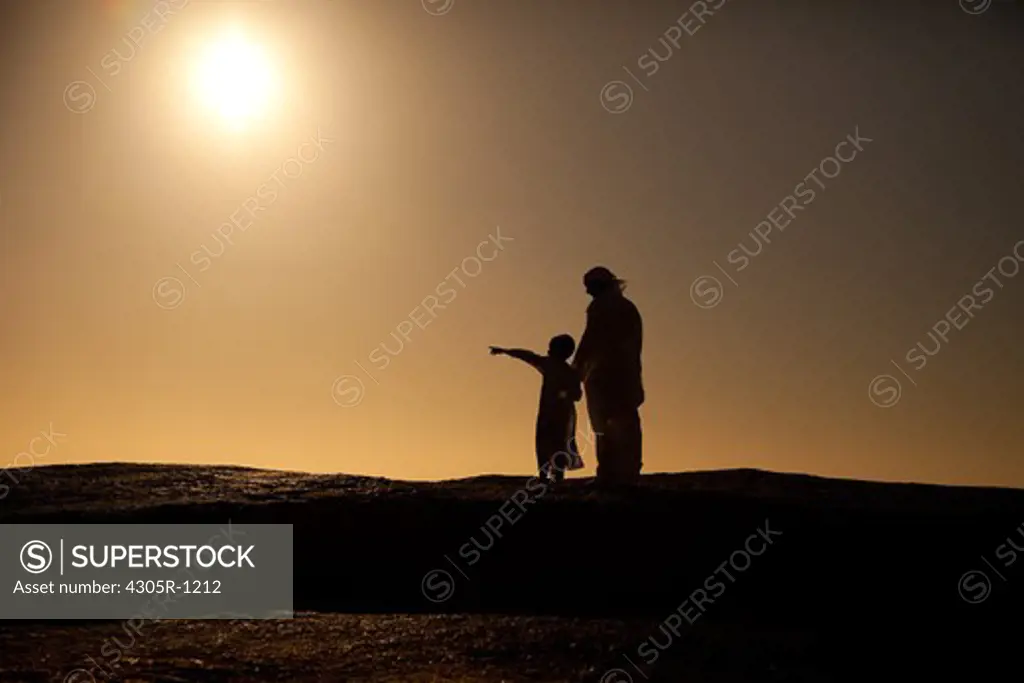 Silhouette of an arab father and son standing on cliff by the beach, boy pointing.