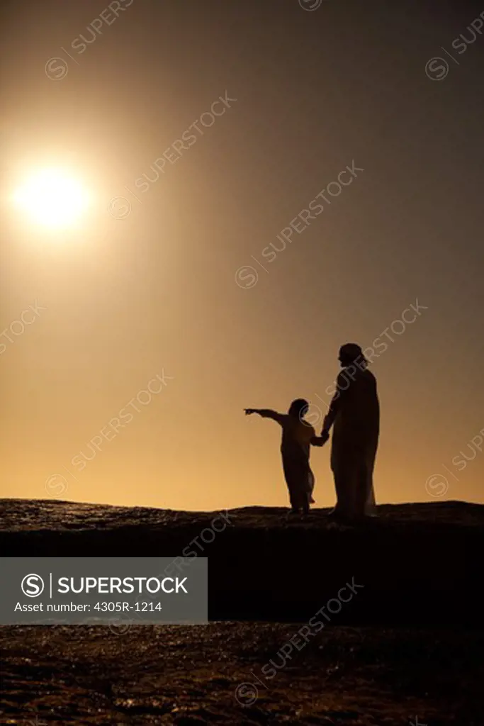 Silhouette of an arab father and son standing on cliff by the beach, boy pointing.