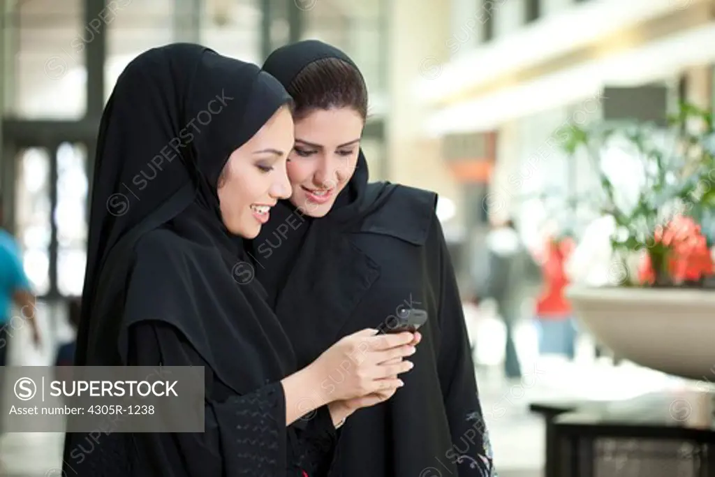 Two young arab women using mobile phone at the shopping mall, smiling.