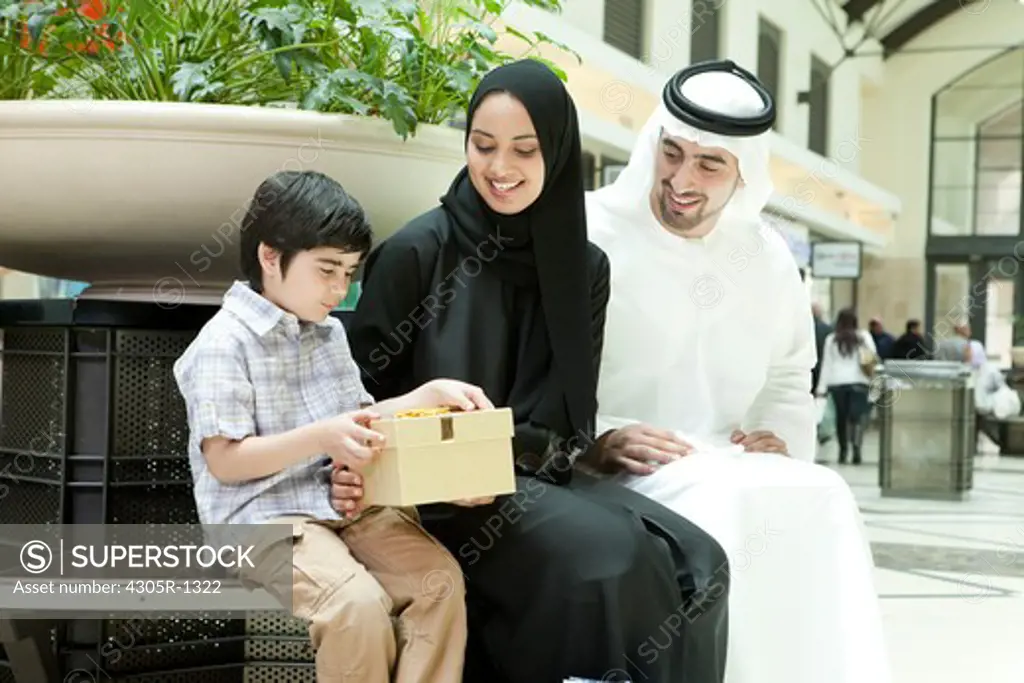 Arab family sitting at the shopping mall, parents giving gift to their son.