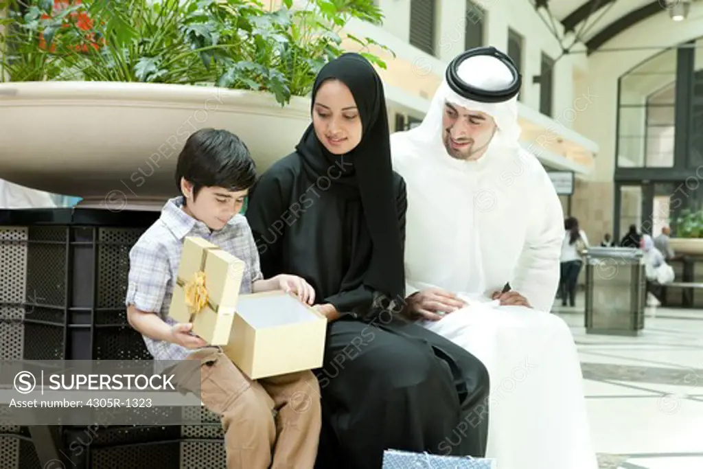 Arab family sitting at the shopping mall, parents giving gift to their son.