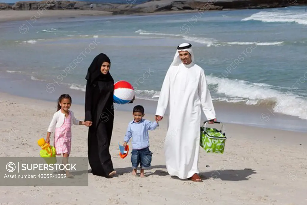 Arab family holding hands while walking at the beach, smiling.