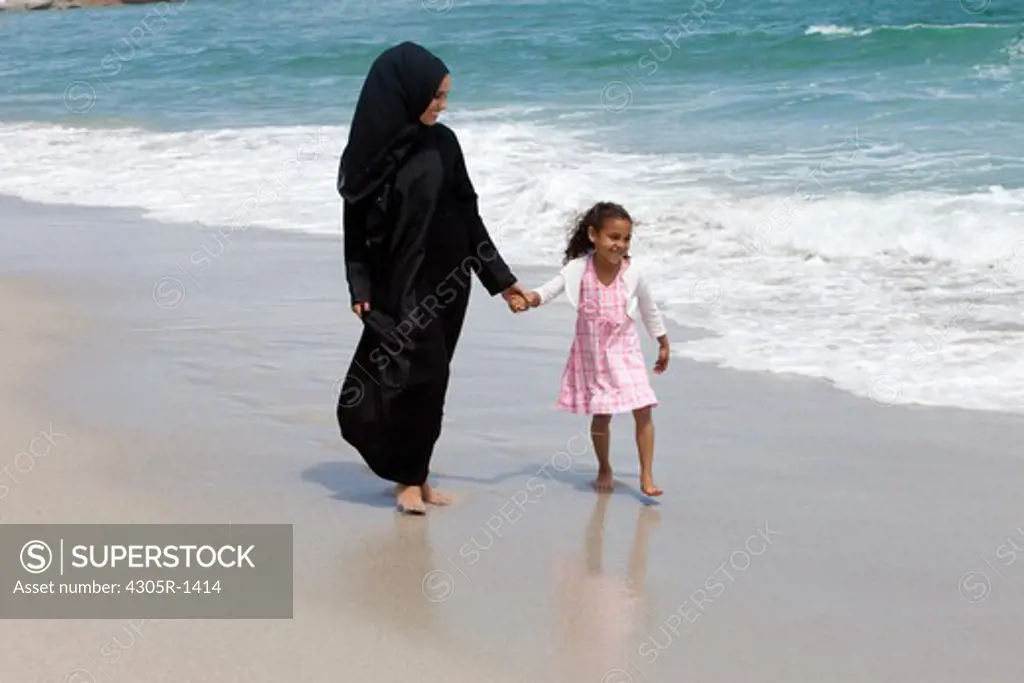 Arab mother and daughter holding hands while walking at the beach.