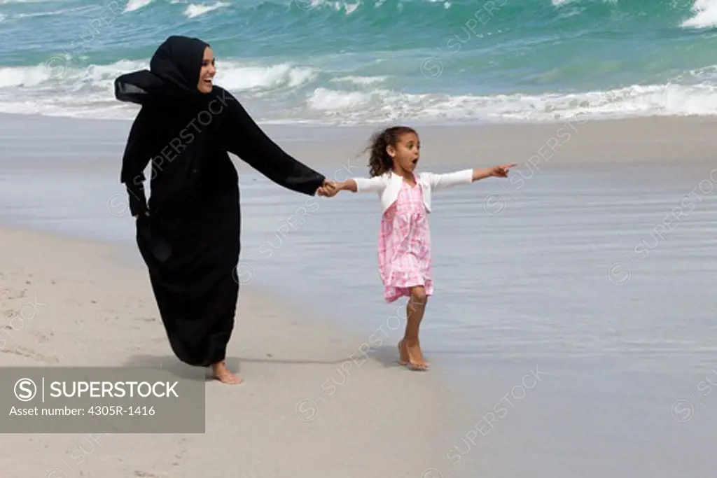 Arab mother and daughter holding hands while walking at the beach, girl pointing.