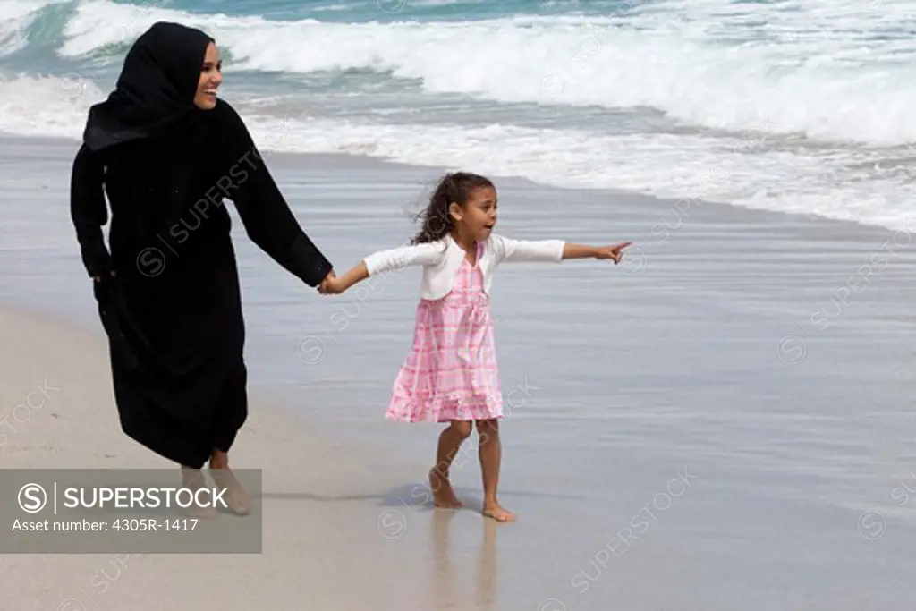 Arab mother and daughter holding hands while walking at the beach, girl pointing.
