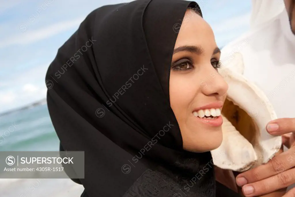 Arab couple at the beach, woman listening to seashell.