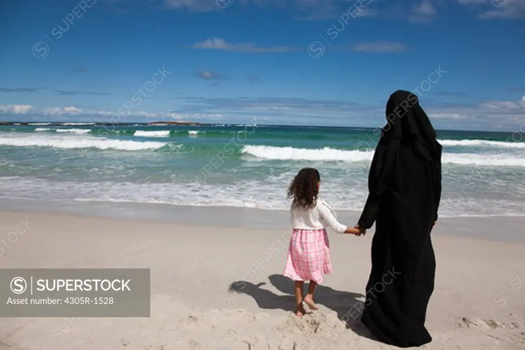 Rear view of arab mother and daughter holding hands at the beach.