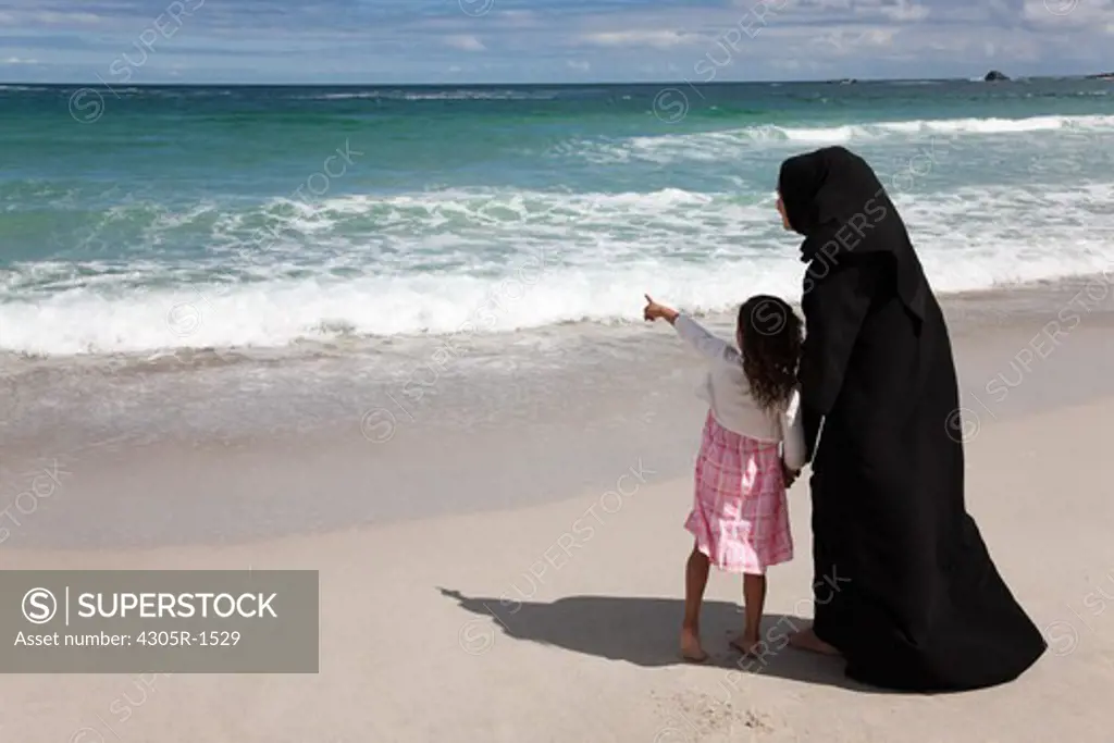 Rear view of arab mother and daughter holding hands at the beach, pointing.