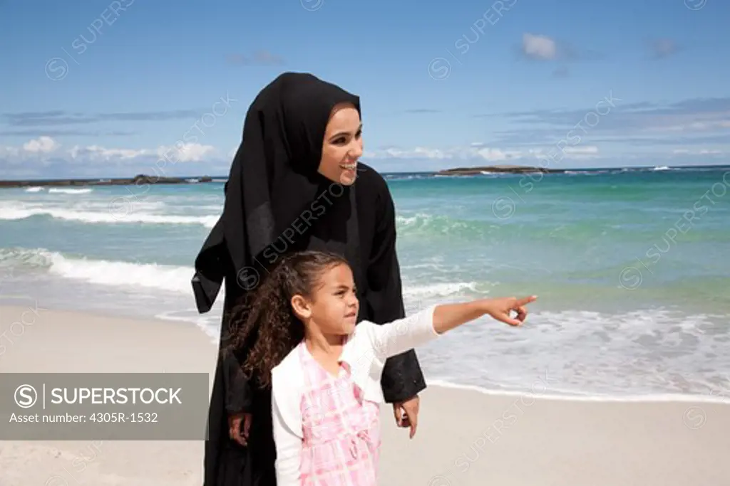 Arab mother and daughter holding hands at the beach, pointing.