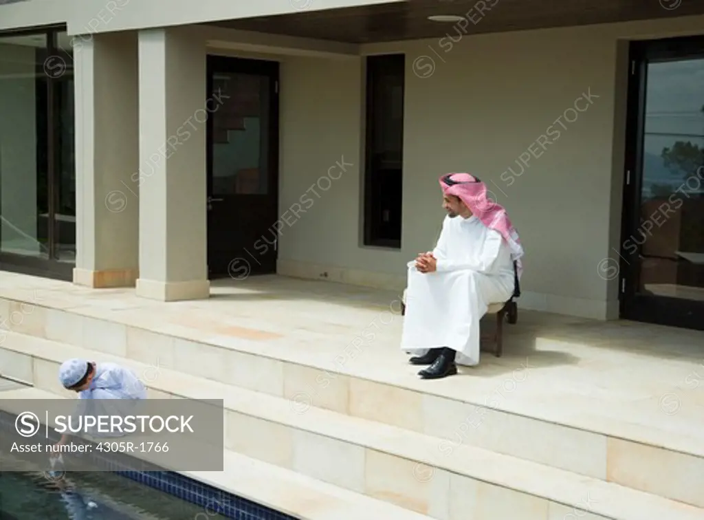 Arab father watching his son, boy playing paper boat at the pool side.