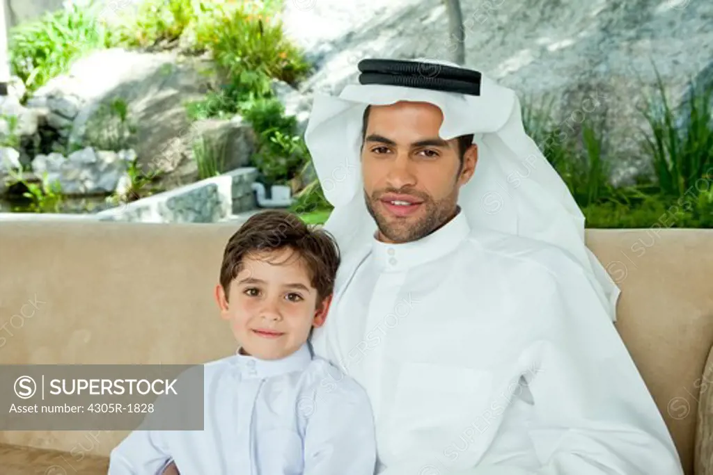 Portrait of an arab father and son sitting on sofa, smiling.