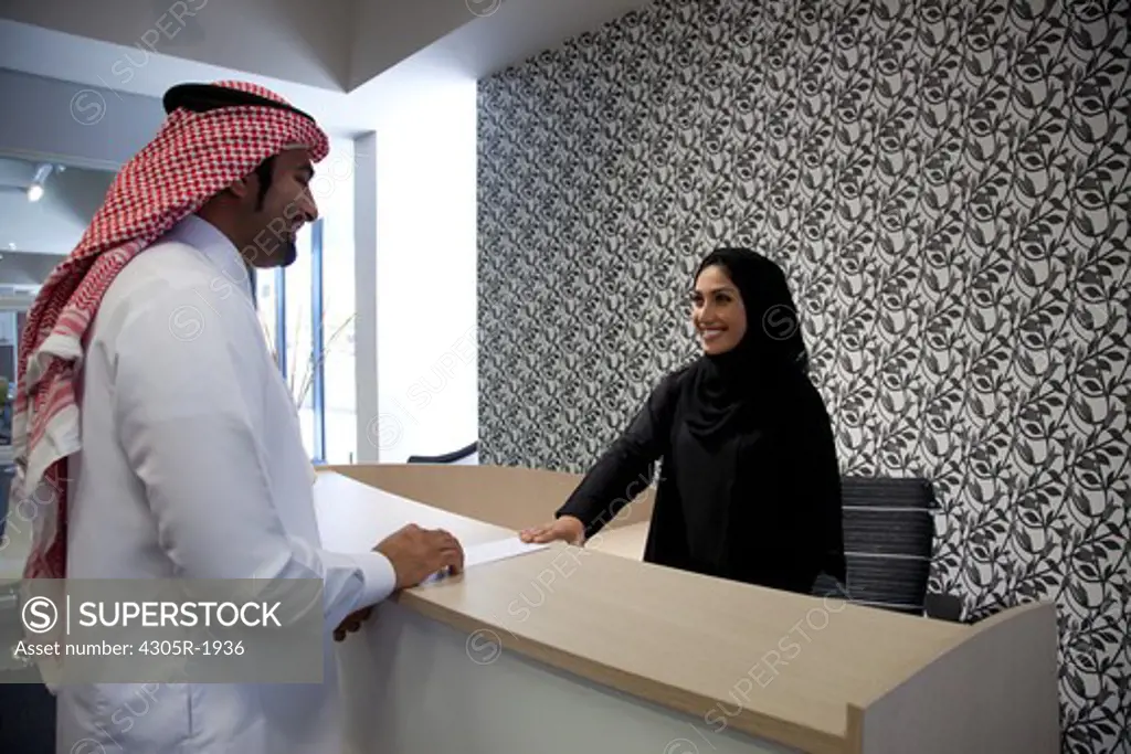 Arab receptionist handing over a document to an arab man, smiling.