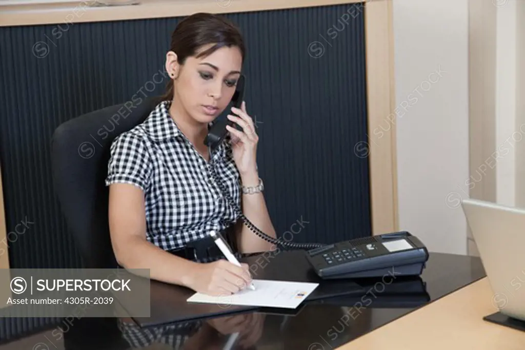 Businesswoman using telephone and writing on notepad at her desk.