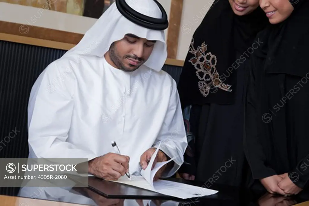Arab businessman signing a document, businesswomen standing on the side.