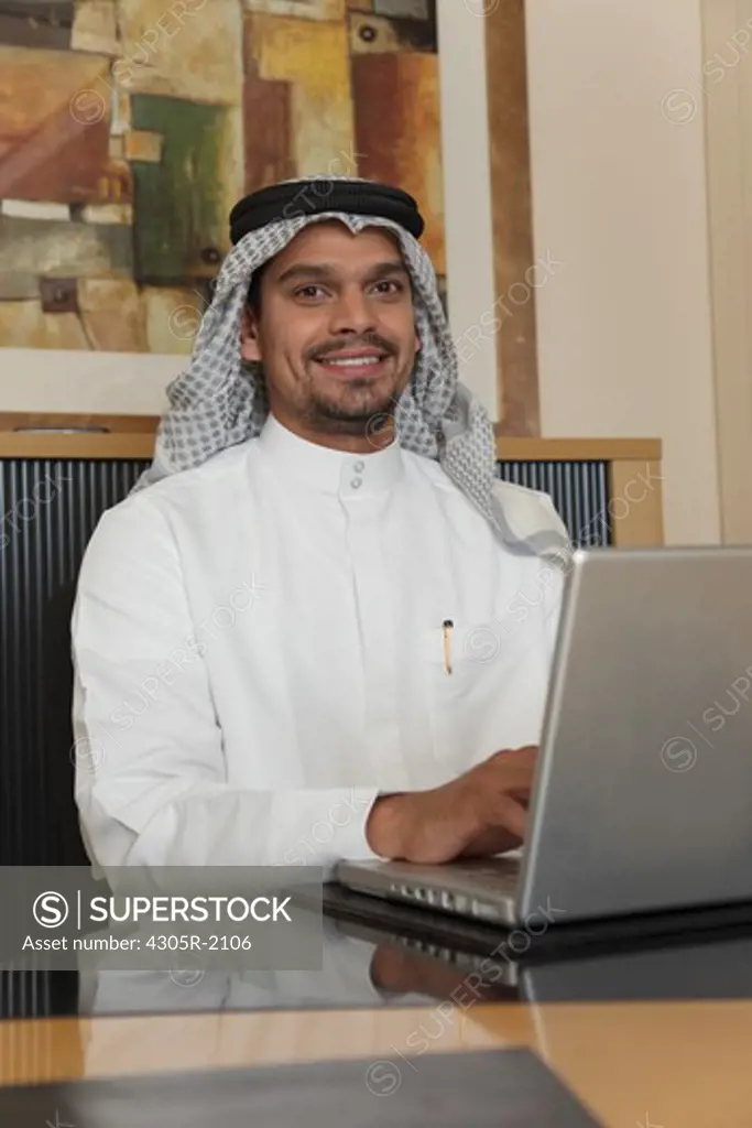 Arab businessman using laptop at desk, smiling.