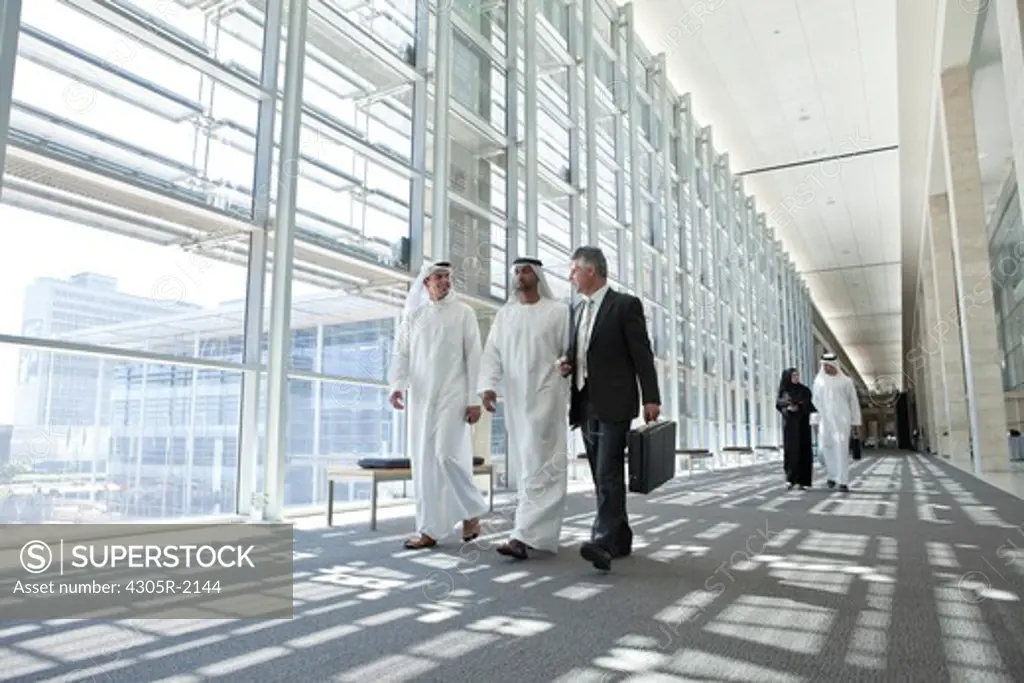 Three businessmen talking in office hallway, arab business man and businesswoman walking in background.