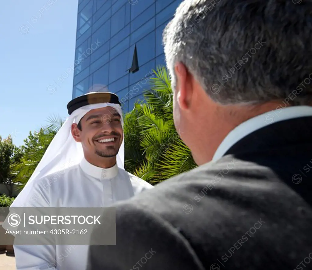 Arab businessman and western businessman in front of building, smiling.
