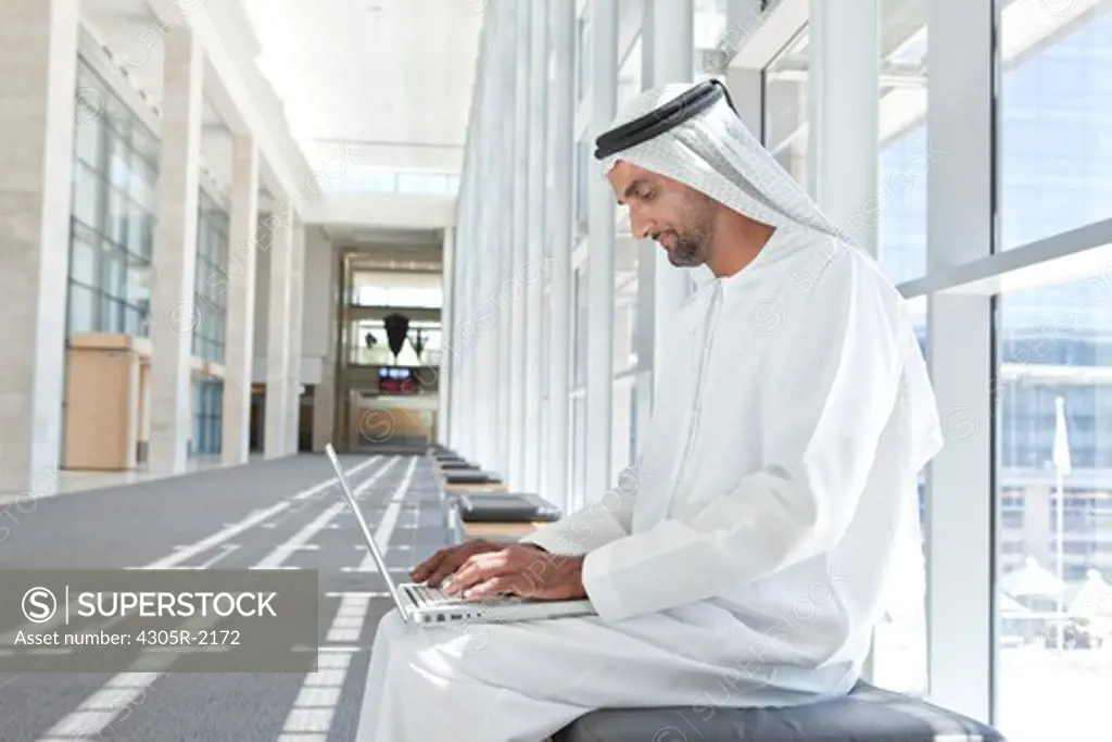 Arab businessman with laptop, sitting on bench on office hallway.