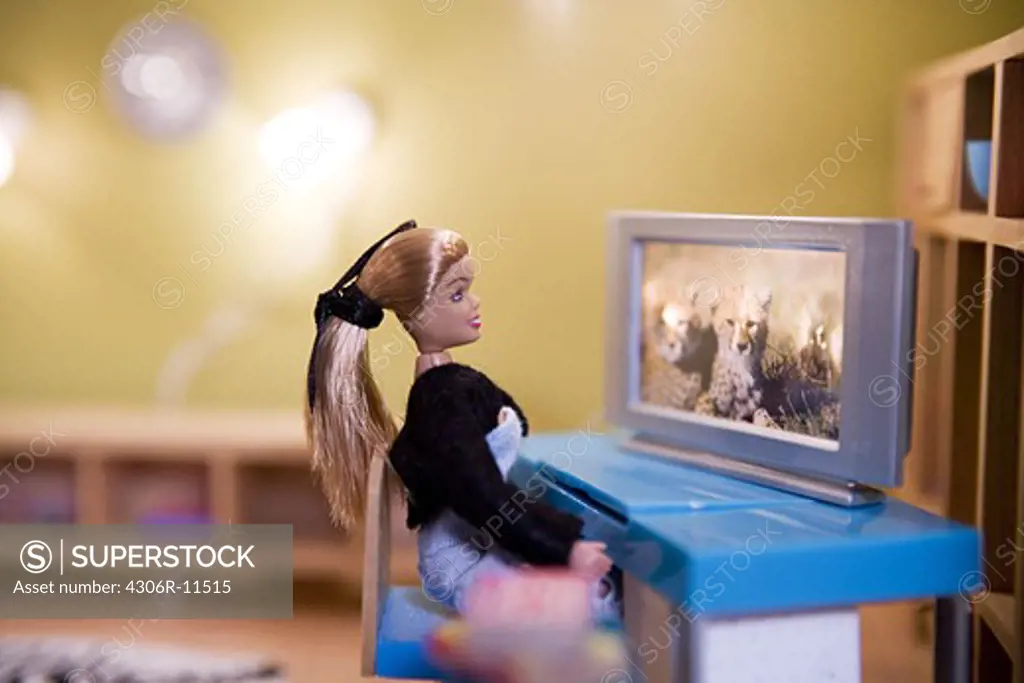A barbie sitting in front of a computer in a dollhouse.
