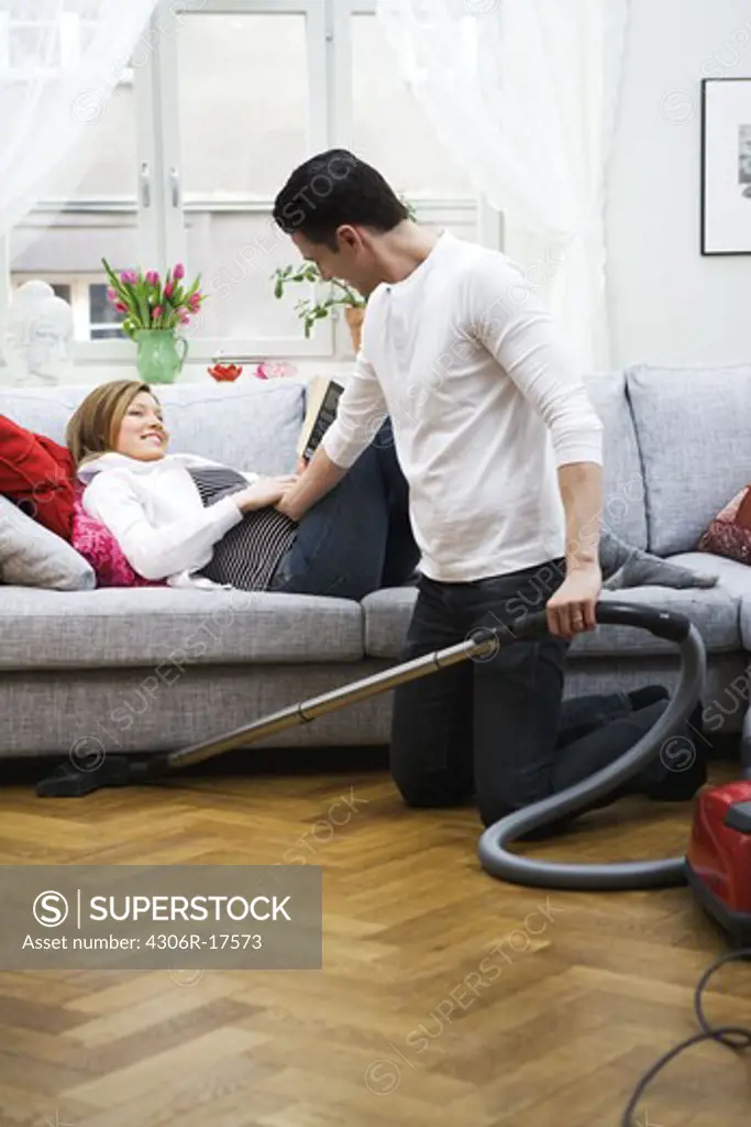 A pregnant woman lying on a couch and a man vacuuming, Sweden.