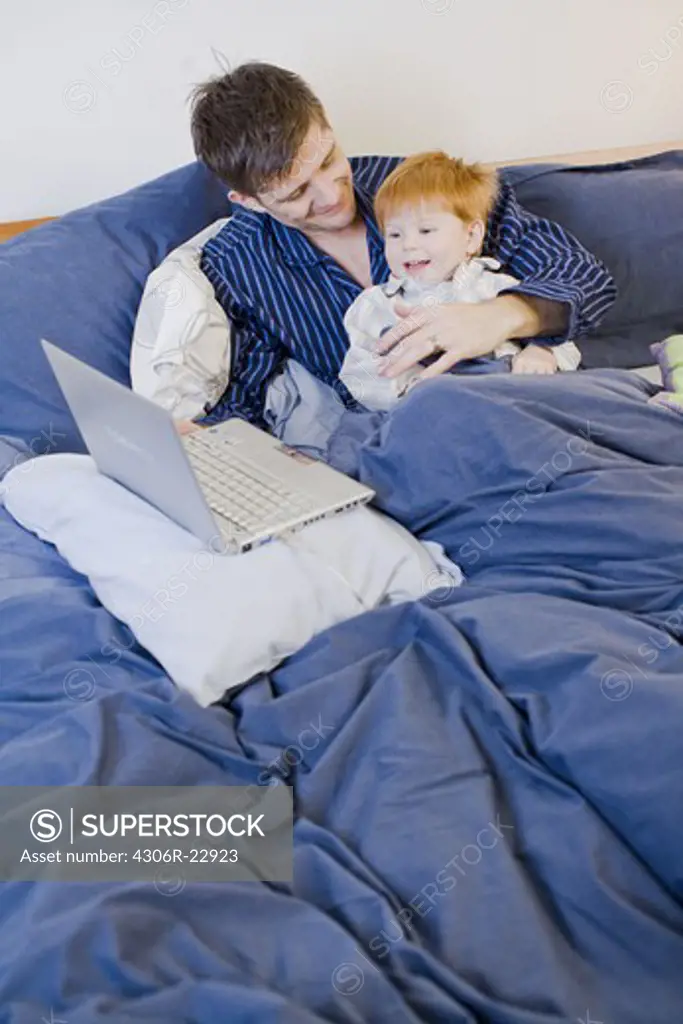 A father and son using a laptop in bed, Sweden.