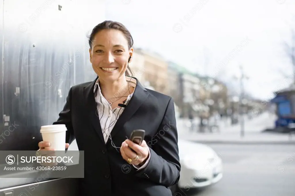 Portrait of cheerful businesswoman with takeaway coffee in one hand and mobile phone in another