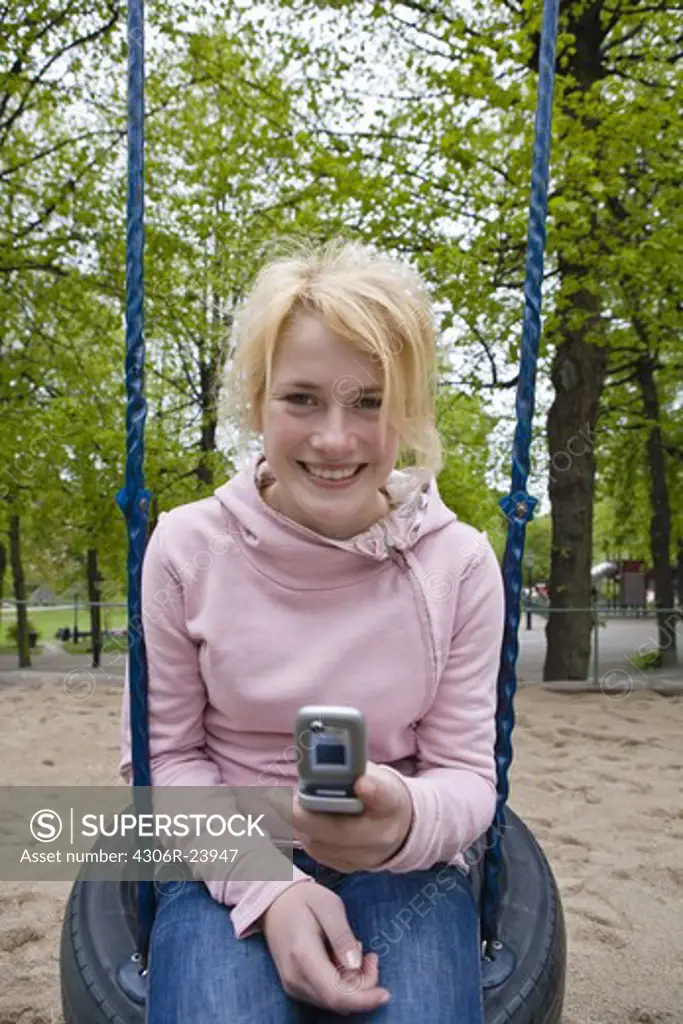 Portrait of smiling teenage girl sitting on tire swing and holding mobile phone