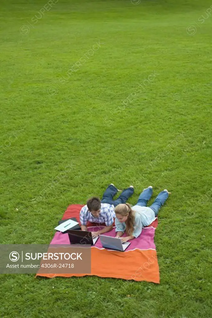 Mid adult couple lying on lawn in park and using laptops