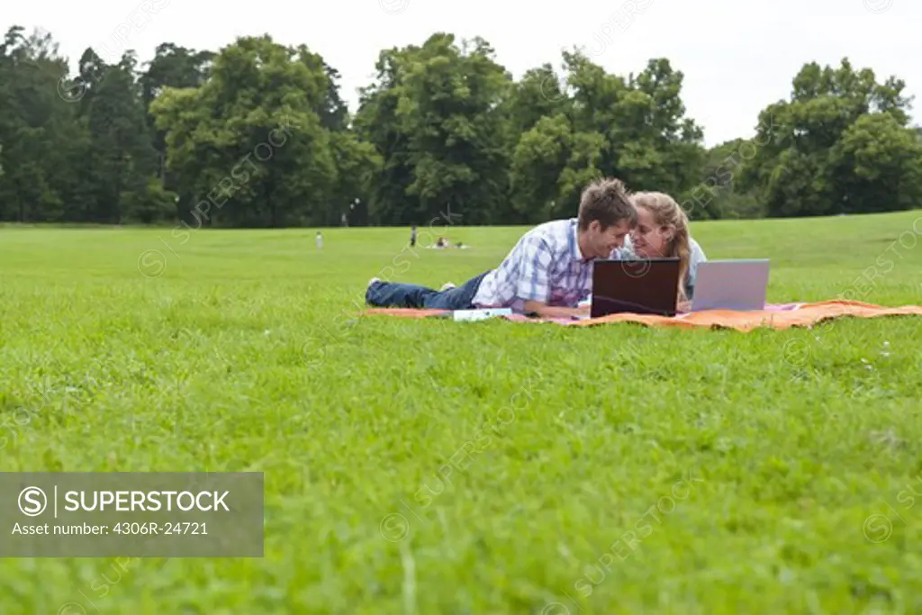 Mid adult couple lying on blanket in park with laptops