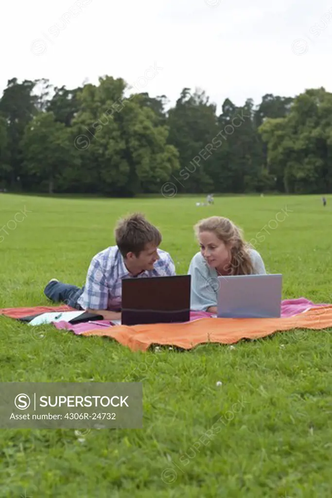 Mid adult couple lying on blanket in park and using laptops