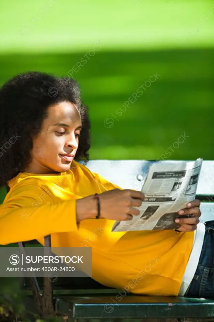 Young man with afro hair  reading newspaper on bench