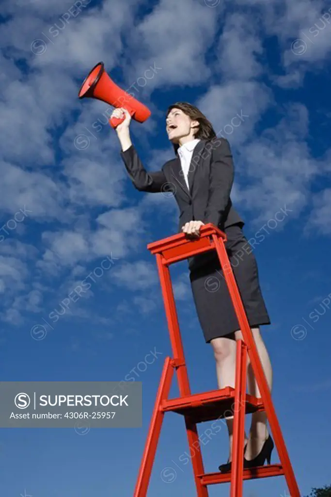 Businesswoman  standing on top of red ladder against blue sky and shouting through red megaphone