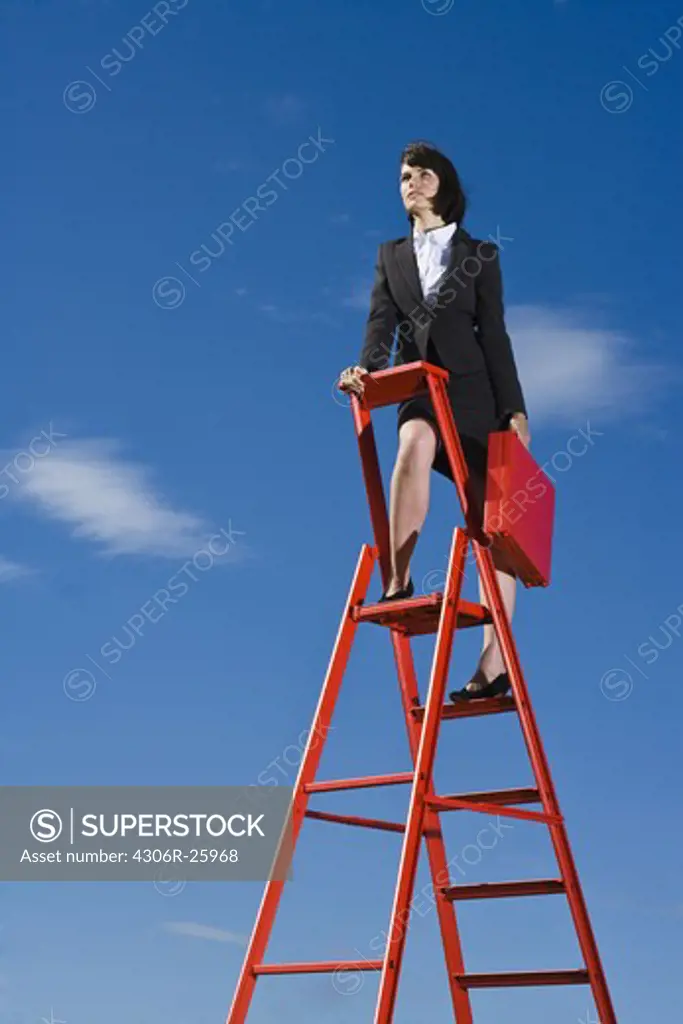 Businesswoman with red briefcase standing on top of red ladder against blue sky