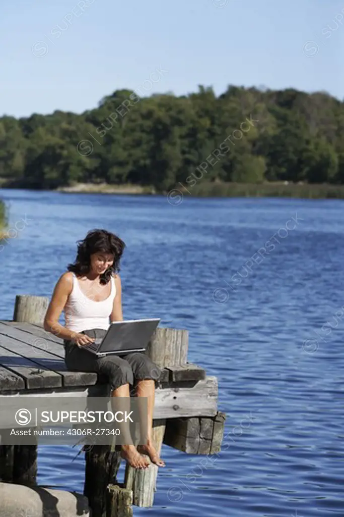 A woman with a laptop on a jetty by a lake, Sweden.