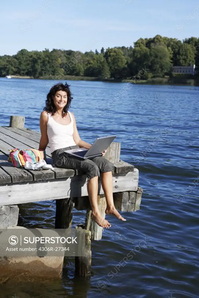 A woman with a laptop on a jetty by a lake, Sweden.