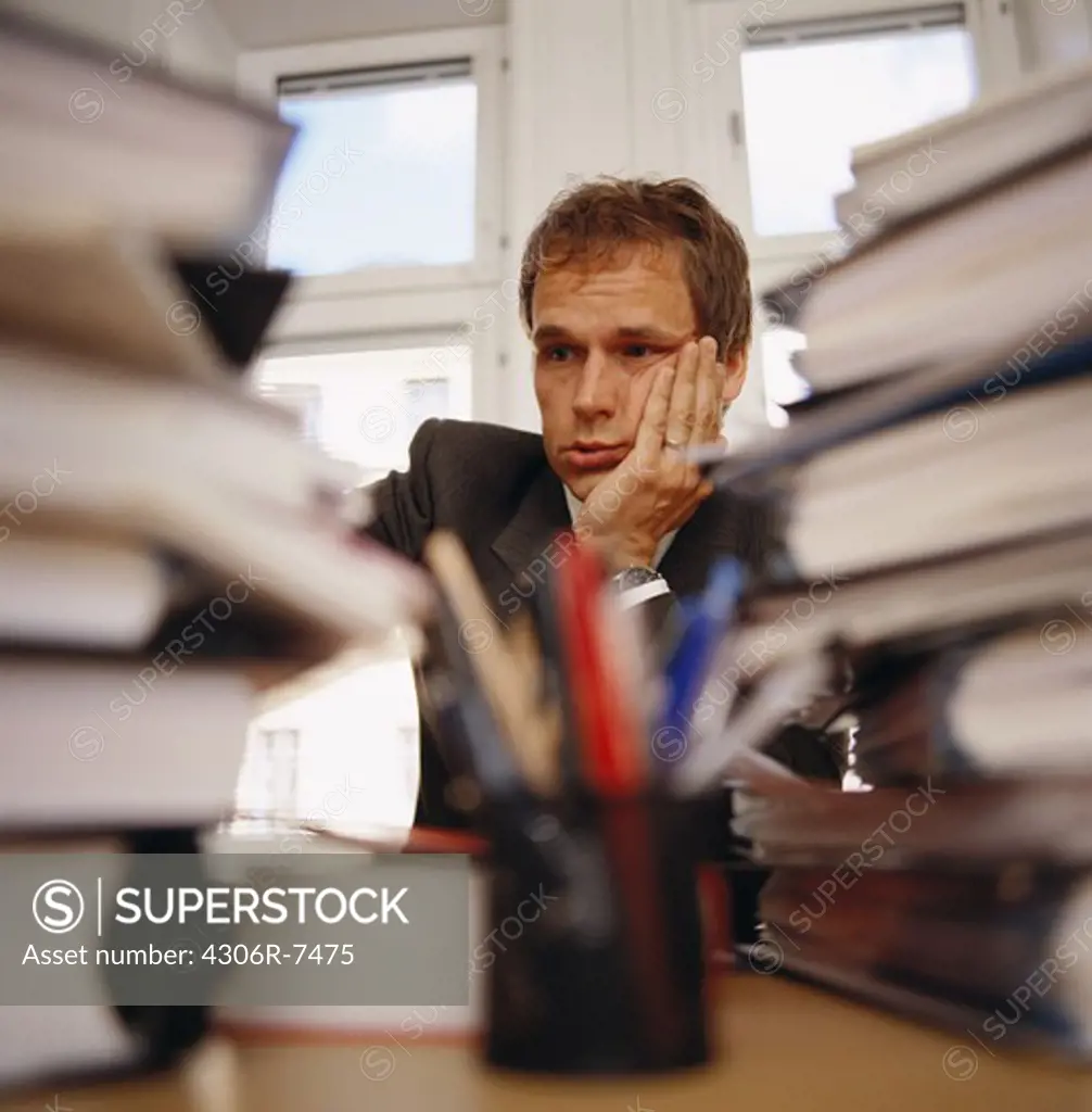 Annoyed businessman sitting with hand on chin behind stack of files