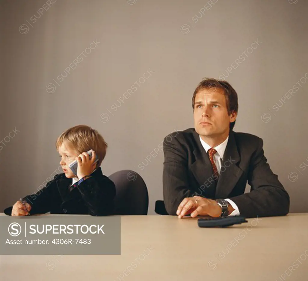 Boy sitting at desk beside annoyed businessman, talking on mobile phone