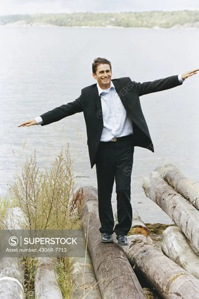 Businessman balancing on logs beside sea with arms out, smiling