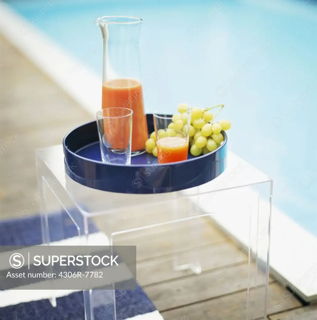 Tray with juice and grapes on glass table beside pool