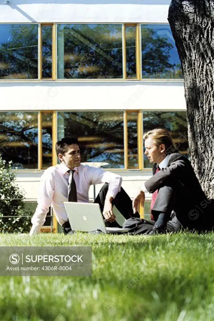 Two businessmen sitting under tree and using laptop in garden
